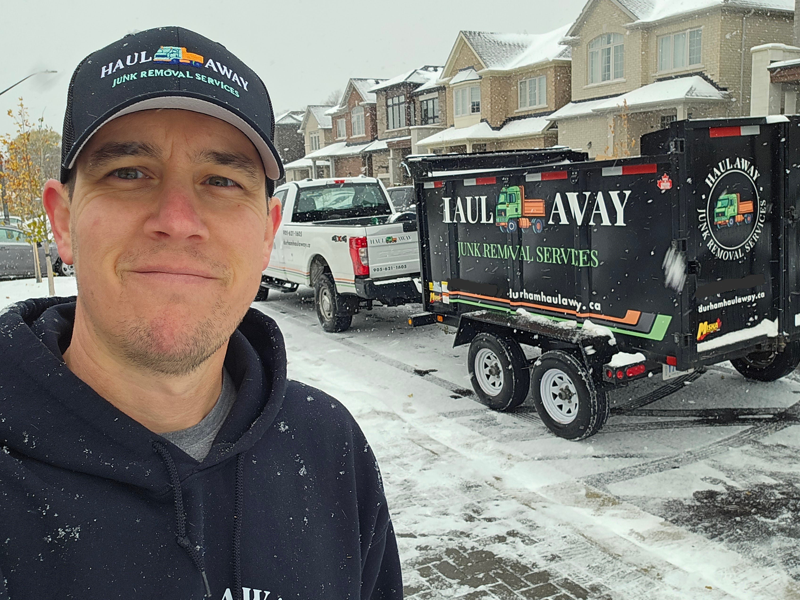 A man stands smiling in the foreground, wearing a black hoodie and cap with the logo 'Haul Away Junk Removal Services'. In the background, a snow-covered street features a truck and a trailer with the same logo parked in front of residential houses.