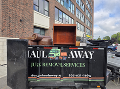 A junk removal trailer filled with various discarded items, including a sofa and a wooden cabinet, parked outside a brick building.